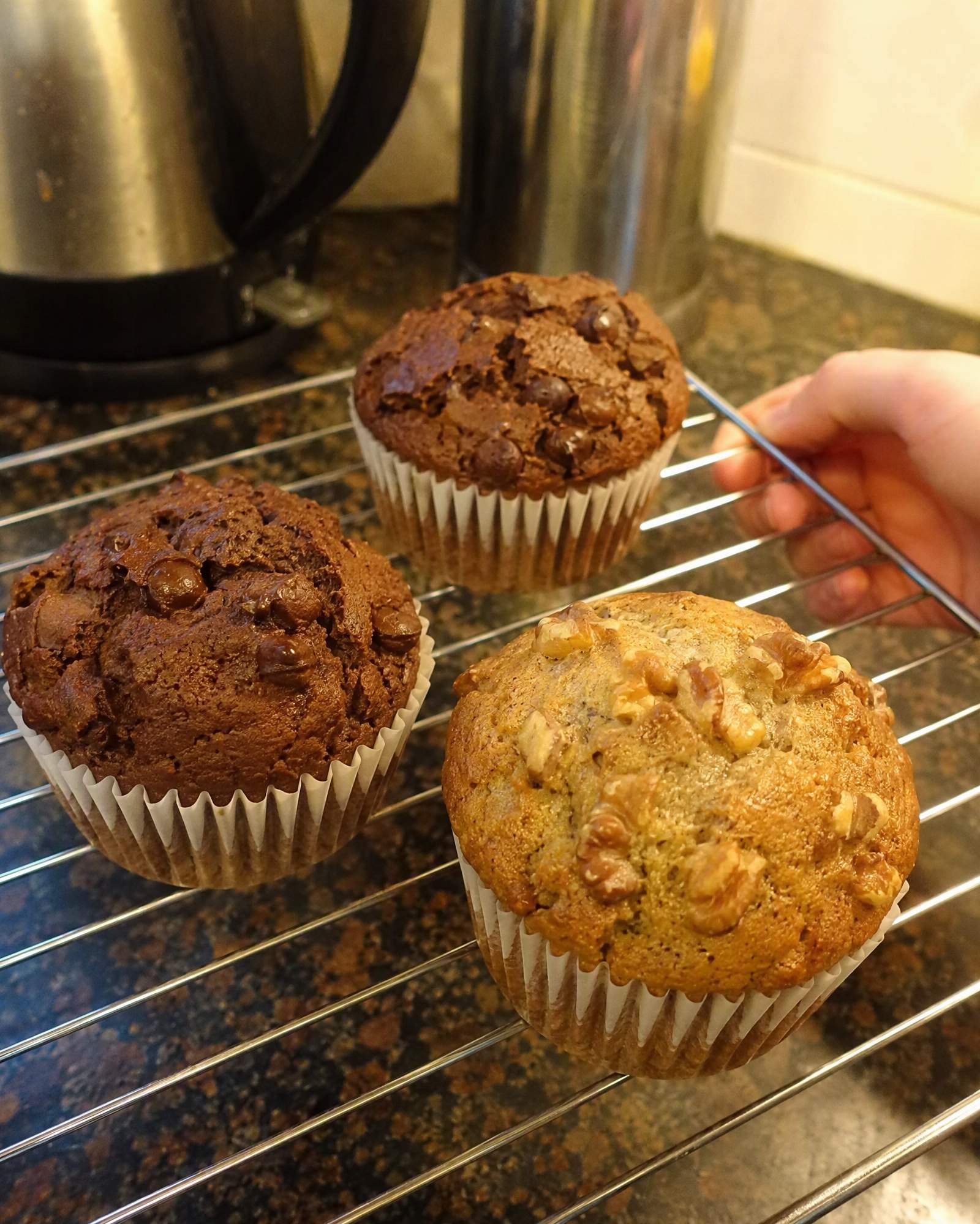 A banana walnut muffin with a crisp, nut-studded top beside two chocolate muffins on a cooling rack