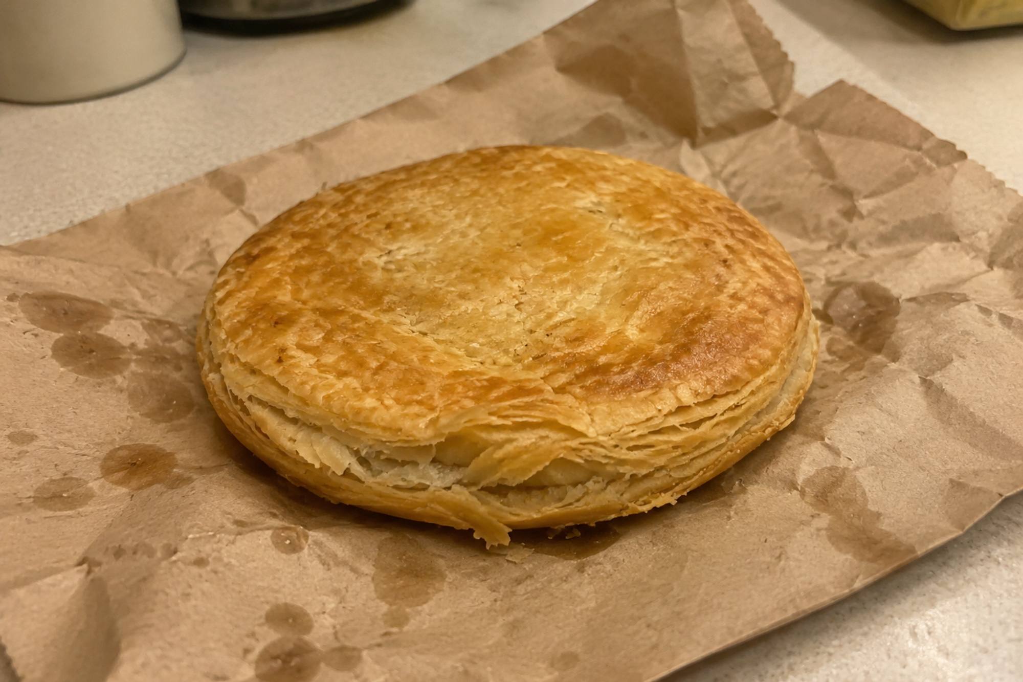 A single flat, round, golden puff pastry chicken pie sitting on a crumpled brown paper bag
