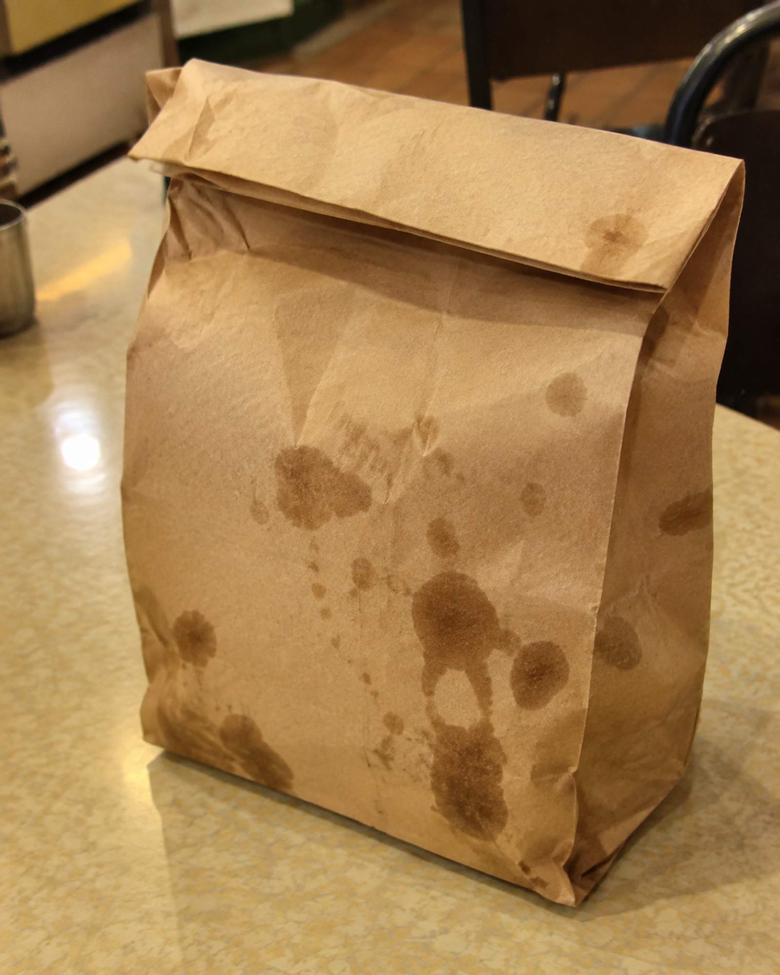 A brown paper bag, top folded over, with grease spots soaking through, standing on a formica table
