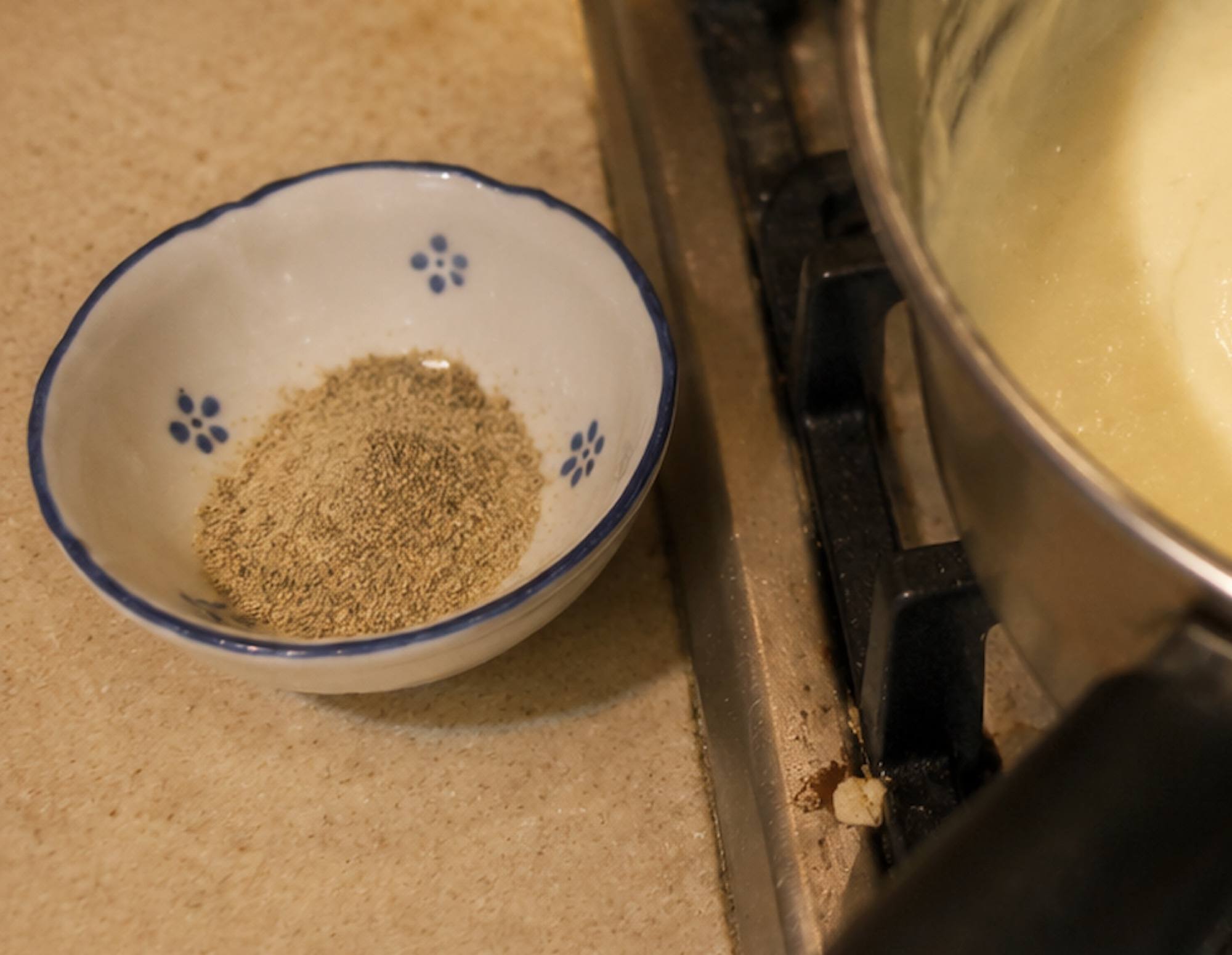 A small blue and white bowl of ground white pepper beside a saucepan of white sauce