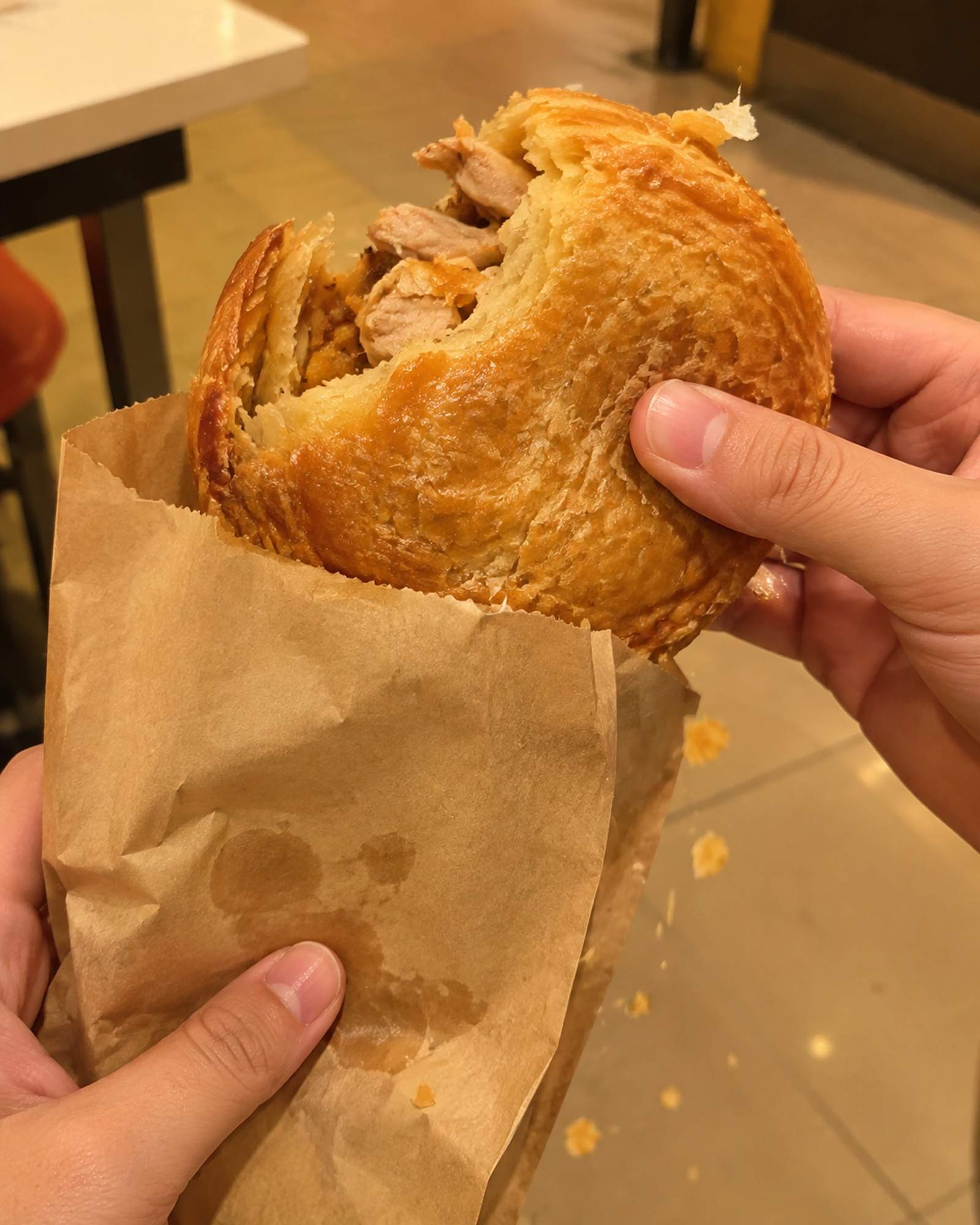 Two hands holding a warm flat puff pastry chicken pie half out of a brown paper bag