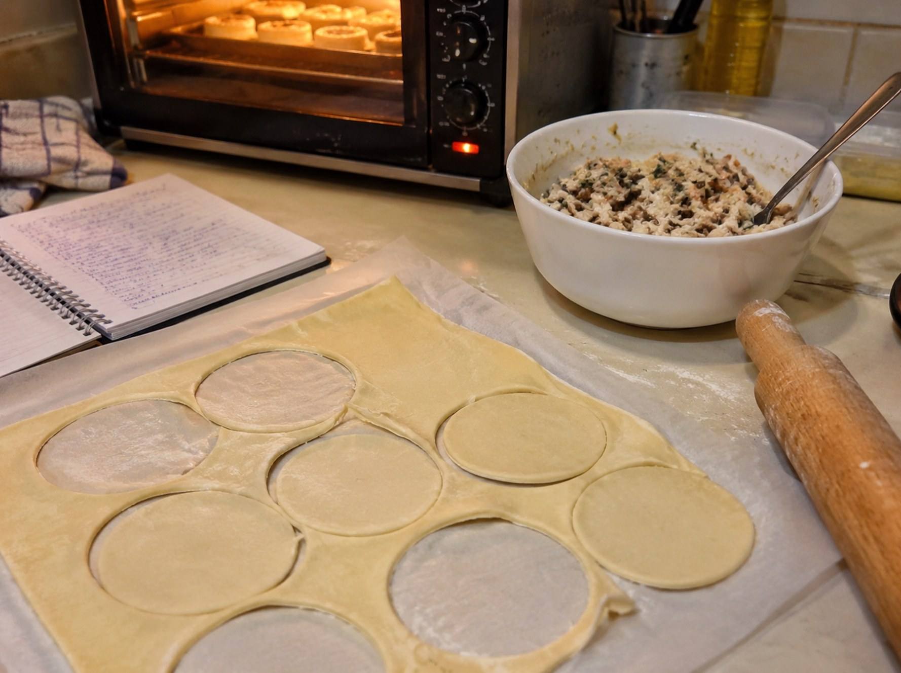 A home kitchen counter mid-bake: pastry circles cut from a sheet, a bowl of filling, a notebook and a glowing oven