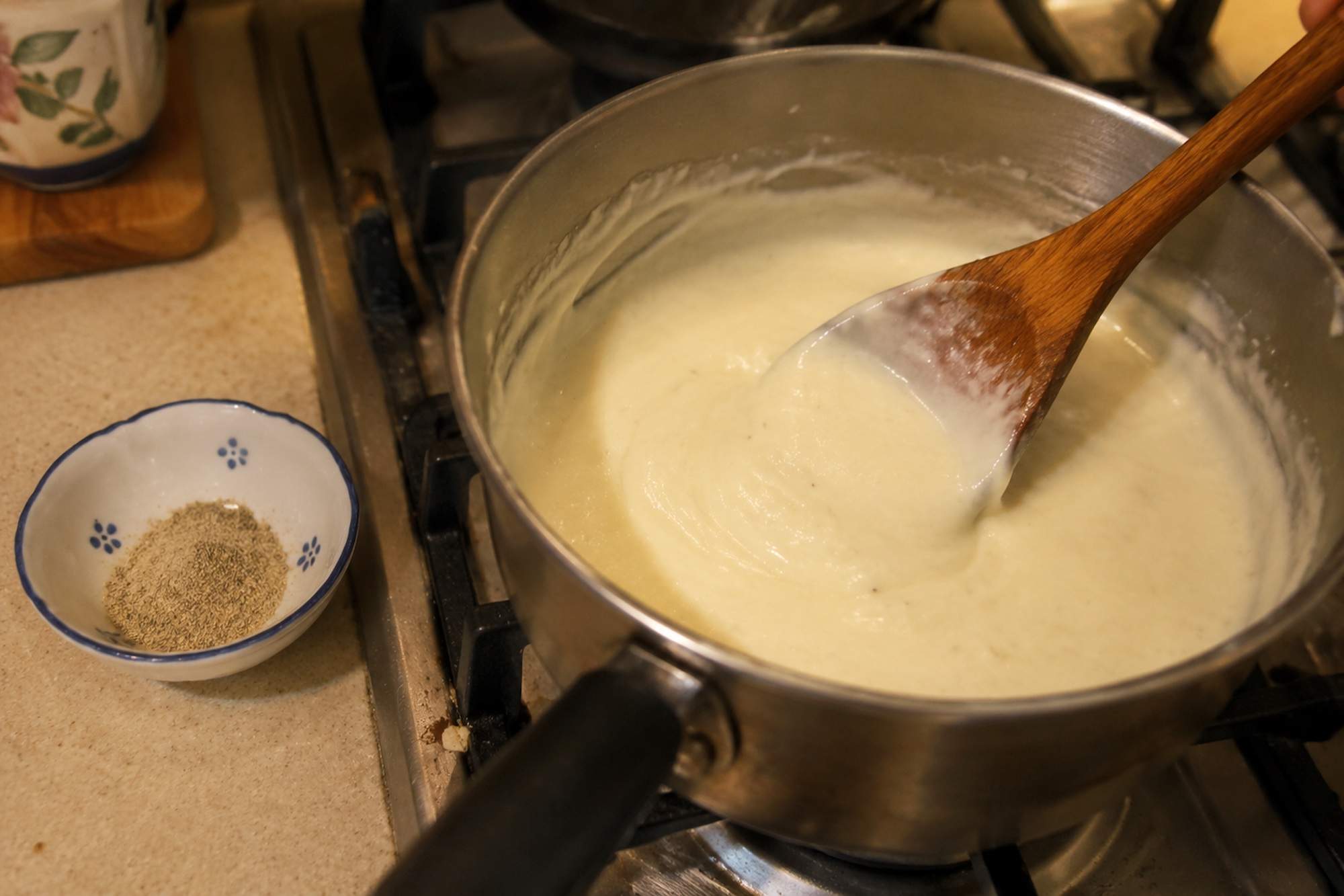 A saucepan of thick white bechamel sauce being stirred with a wooden spoon, with a small bowl of ground white pepper beside it